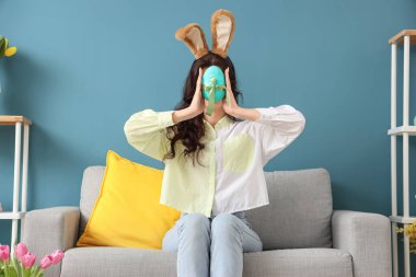 Tavşan kulaklı genç bir kadın. Paskalya yumurtası evde, kanepede oturuyor.