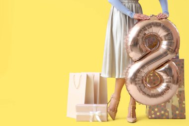 8 numaralı figür şeklinde balonu olan genç bir kadın, sarı arka planda hediye ve alışveriş torbaları. Uluslararası Kadınlar Günü