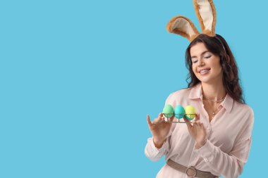 Mavi arka planda boyanmış Paskalya yumurtaları olan tavşan kulaklı güzel bir kadın.
