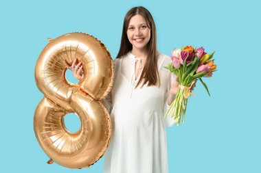 Güzel, genç, hamile bir kadın. Mavi arka planda, elinde bir buket lale ve altın renkli balon var. Uluslararası Kadınlar Günü