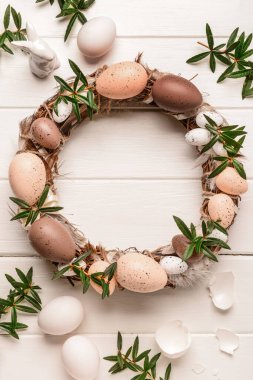 Beyaz ahşap masada yumurta ve çiçeklerle süslenmiş şık bir Paskalya çelengi. Üst görünüm
