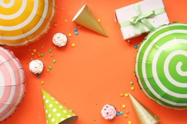 Hediye kutulu balonlar, turuncu arka planda parti şapkaları ve kekler.