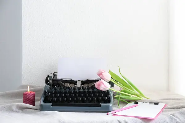Vintage typewriter with tulips, candle and clipboard on cloth near white wall