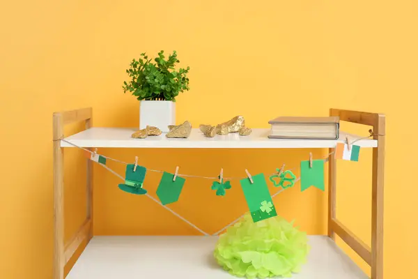 Shelving unit with gold, green pompom and garland for St. Patrick\'s Day celebration near orange wall