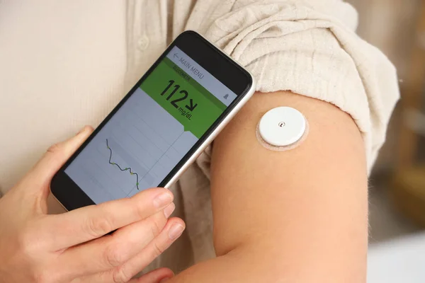 Diabetic woman with glucose sensor using mobile phone for measuring blood sugar level in bedroom, closeup