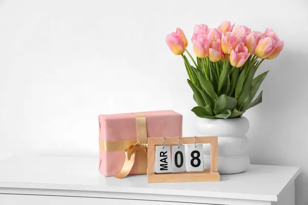 Calendar with date of International Women\'s Day, tulips and gift box on table near light wall