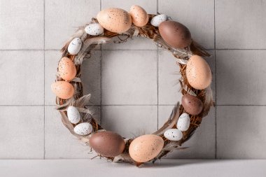 Beyaz duvarın yanındaki masada yumurta ve tüylerden yapılmış Paskalya çelengi.