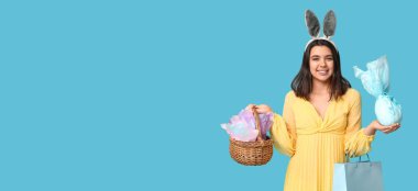 Tavşan kulaklı güzel genç kadın, Paskalya hediyeleri, sepet ve alışveriş çantası açık mavi arka planda mesaj için yer var.