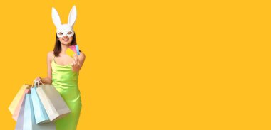 Tavşanlı güzel bir genç kadın elinde alışveriş çantaları ve kredi kartlarıyla sarı arka planda mesajlaşmak için yer açıyor. Paskalya indirimi afişi