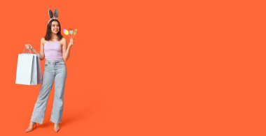 Tavşan kulaklı, dekorlu ve turuncu arka planda alışveriş torbaları olan güzel bir genç kadın. Paskalya indirimi