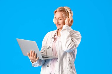 Kulaklıklı yetişkin bir kadın programcı mavi arka planda dizüstü bilgisayarla çalışıyor.