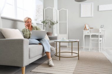 Evde koltukta cep telefonuyla konuşan, dizüstü bilgisayarı olan olgun bir programcı.