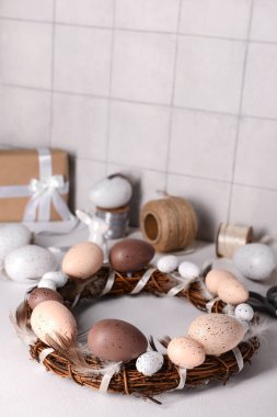 Yumurta ve tüylerden yapılmış Paskalya çelengi. Beyaz duvarın yanındaki masada erzak var.