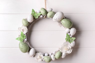 Beyaz tahta duvarda asılı, çiçeklerden, yumurtalardan ve kelebeklerden yapılmış Paskalya çelengi.