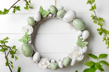 Beyaz ahşap duvarda asılı dalları olan çiçeklerden, yumurtalardan ve kelebeklerden oluşan Paskalya çelengi.