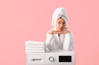 Bornozlu düşünceli genç bir kadın. Pembe arka planda çamaşır makinesinin üstüne yığılmış temiz giysiler.