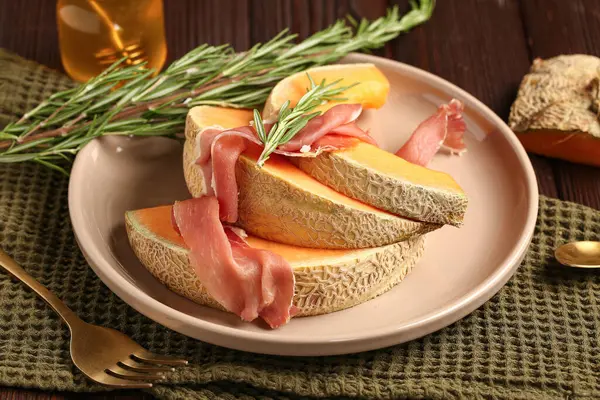 Plate with tasty melon, prosciutto and rosemary on wooden table, closeup