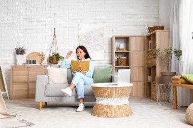 Şık kapüşonlu güzel Asyalı kadın kanepede oturup evde kitap okuyor.