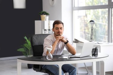 Ofiste oturan laptopu, tableti ve kum saati olan yakışıklı bir genç.