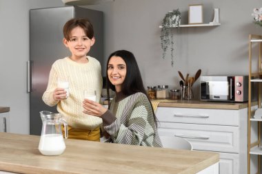 Mutlu küçük çocuk ve annesi mutfakta bir bardak taze sütle masada oturuyorlar.