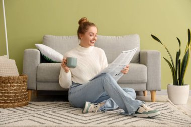 Elinde kahve fincanı olan genç bir kadın evde gazete okuyor.