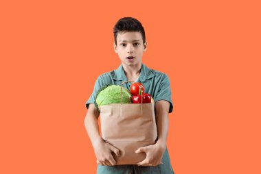 Turuncu arka planda farklı sebzelerle dolu kese kağıdı tutan sevimli bir çocuk.