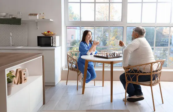 Mature man and nurse with tea playing chess at table in kitchen
