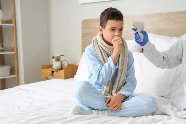 Doctor with infrared thermometer measuring temperature of sick little boy in bedroom