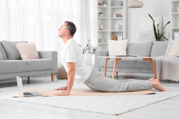 Mature man with laptop doing yoga on mat at home
