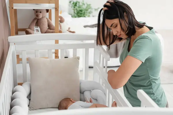 Young woman with her baby in crib suffering from postnatal depression at home