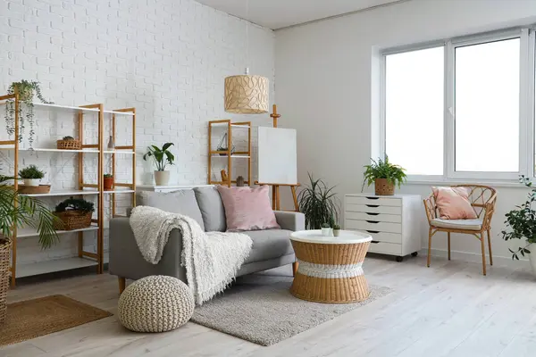 Loft style interior of light living room with cozy sofa, chest drawer, table, houseplants, shelving units, armchair and easel