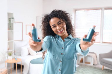 Smiling young African-American woman with asthma inhalers in bedroom clipart