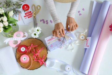 Hediye kutusu olan kadın eller, hafif arka planda paketleme malzemeleri ve çiçekler. Uluslararası Kadınlar Günü