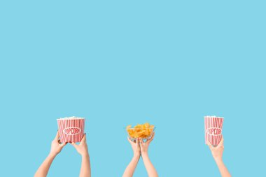 Mavi arka planda patlamış mısır ve patates cipsi dolu kovaları tutan eller.