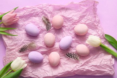 Kırışmış kağıt, boyalı Paskalya yumurtaları, tüyler ve mor arka planda lale çiçekleri.