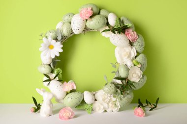 Küçük tavşan, yeşil arka planda çiçekler ve yumurtalarla süslenmiş Paskalya çelengi.