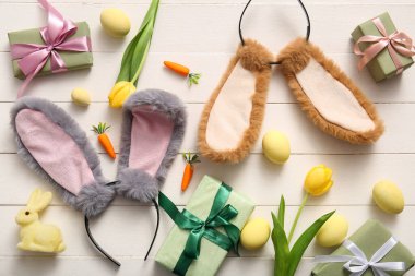 Paskalya yumurtalı tavşan kulakları, havuç, hediye kutuları ve beyaz ahşap arka planda lale çiçekleri.
