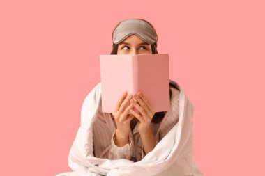 Battaniyeli, uyku maskeli ve pembe arka planda kitap okuyan güzel bir kadın.