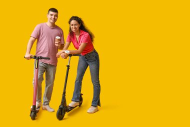 Modern elektrikli scooterları ve sarı arka planda kağıt kahve fincanları olan genç ve güzel bir çift.