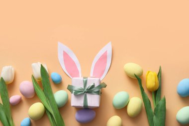 Hediye kutusuyla boyanmış Paskalya yumurtaları, lale çiçekleri ve bej arka planda kağıt tavşan kulakları.