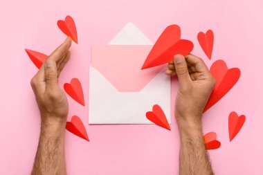 Zarflı erkek eller, boş kart ve pembe arka planda kırmızı kağıt kalpler. Sevgililer Günü kutlaması