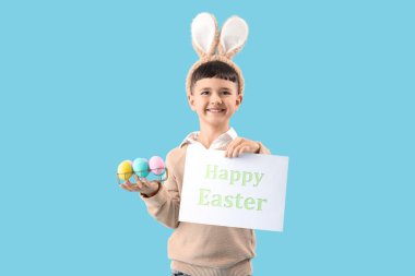 Paskalya yumurtaları ve mavi arka planda hediye kartıyla tavşan kulağı takan şirin bir çocuk.