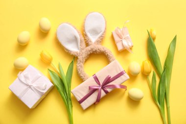 Paskalya yumurtalı tavşan kulakları, lale çiçekleri ve sarı arka planda hediye kutuları.