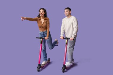 Modern elektrikli scooter kullanan ve mor arka plandaki bir şeyi işaret eden genç ve güzel bir çift.