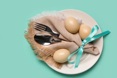 Renkli Paskalya yumurtaları, peçete ve turkuaz arka planda çatal bıçak seti olan şık bir masa.