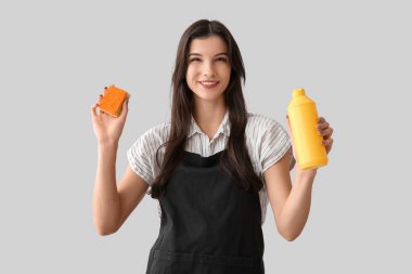 Gri arka planda sünger ve deterjanı olan genç bir kadın.