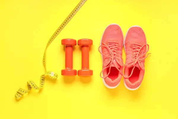 Dumbbells, sportive shoes and measuring tape on yellow background