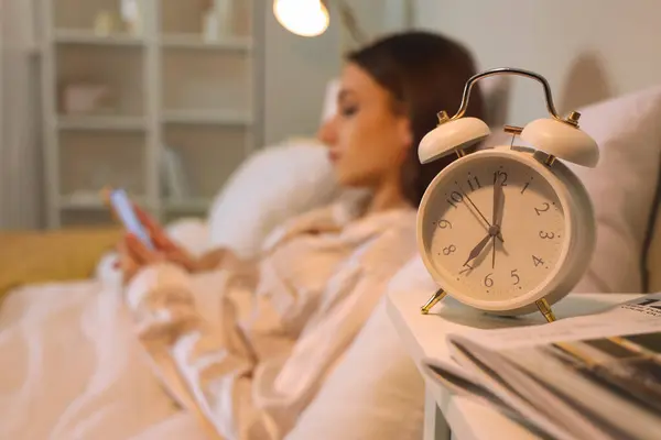 Alarm clock on table of young woman using mobile phone in bedroom at night, closeup