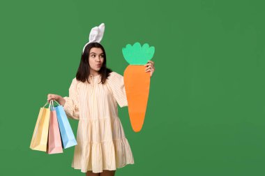 Alışveriş torbaları ve yeşil arka planda kağıt havuçla tavşan kulaklı güzel, genç ve düşünceli bir kadın. Paskalya kutlaması
