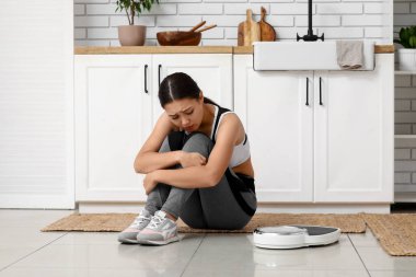 Mutfakta terazinin yanında oturan üzgün, spor giyimli genç bir kadın. Kilo verme konsepti